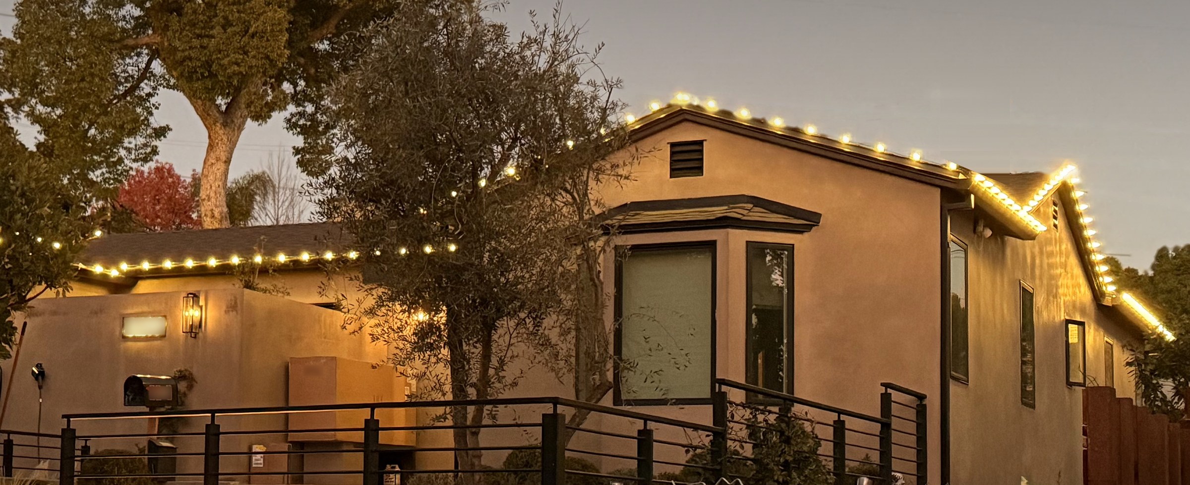 Street view of a stucco home at dusk, its rooflines and front gable outlined in warm white lights