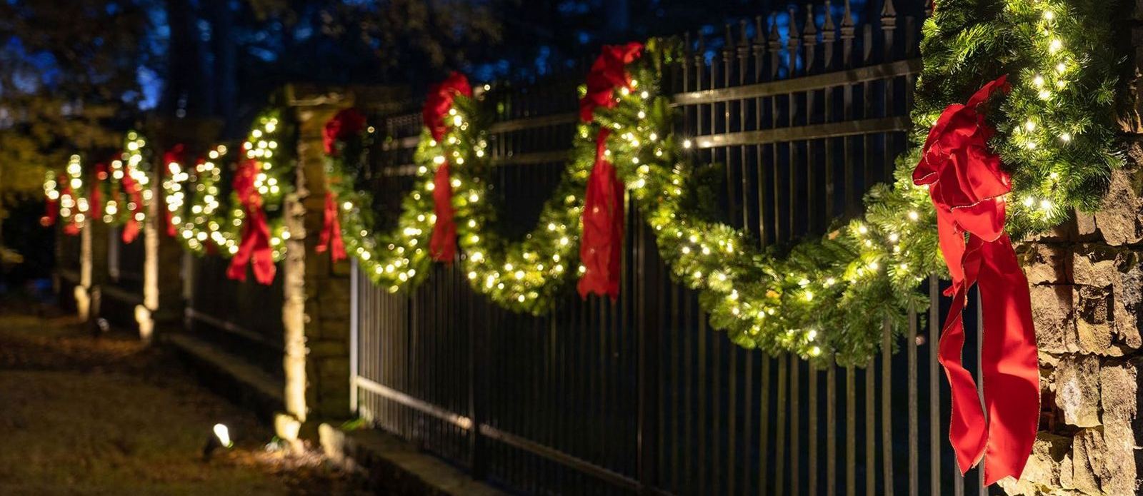 An iron fence lined with lit garland and red bows at night
