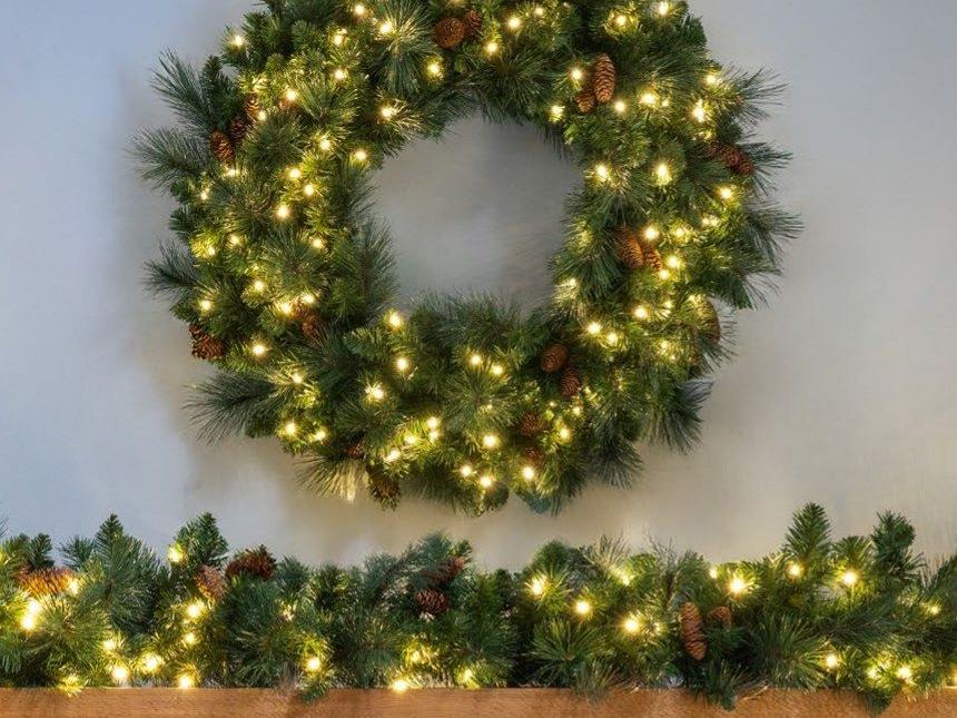 A lit wreath with pine cones above a matching lit garland on a mantel