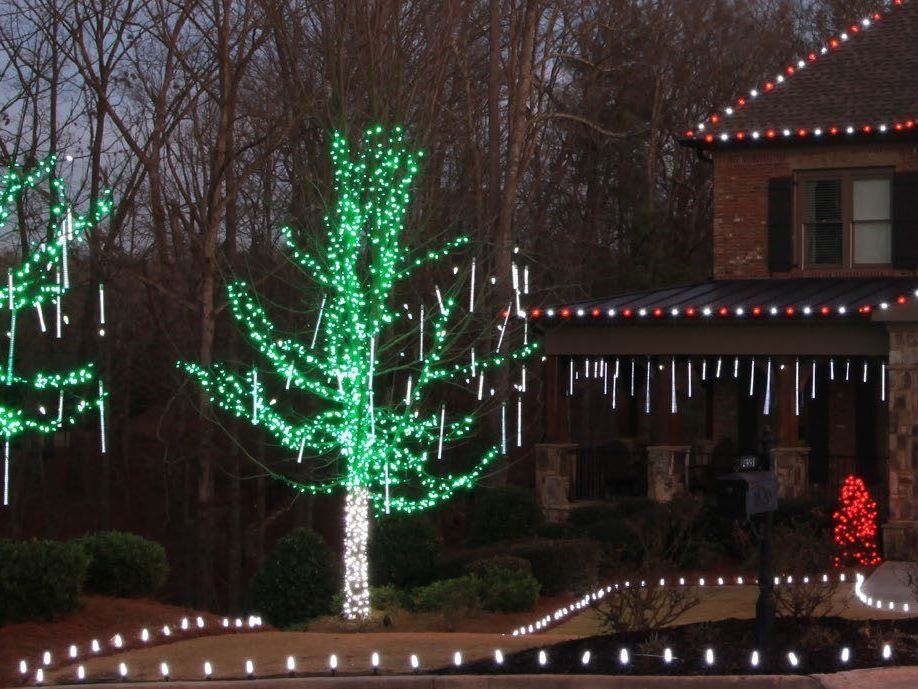 Trees wrapped in green lights with cascading tubes, in front of a lit house